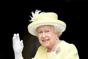 La reina Isabel II dice adiós con la mano. Va vestida de amarillo, traje y sombrero. Guante blanco.