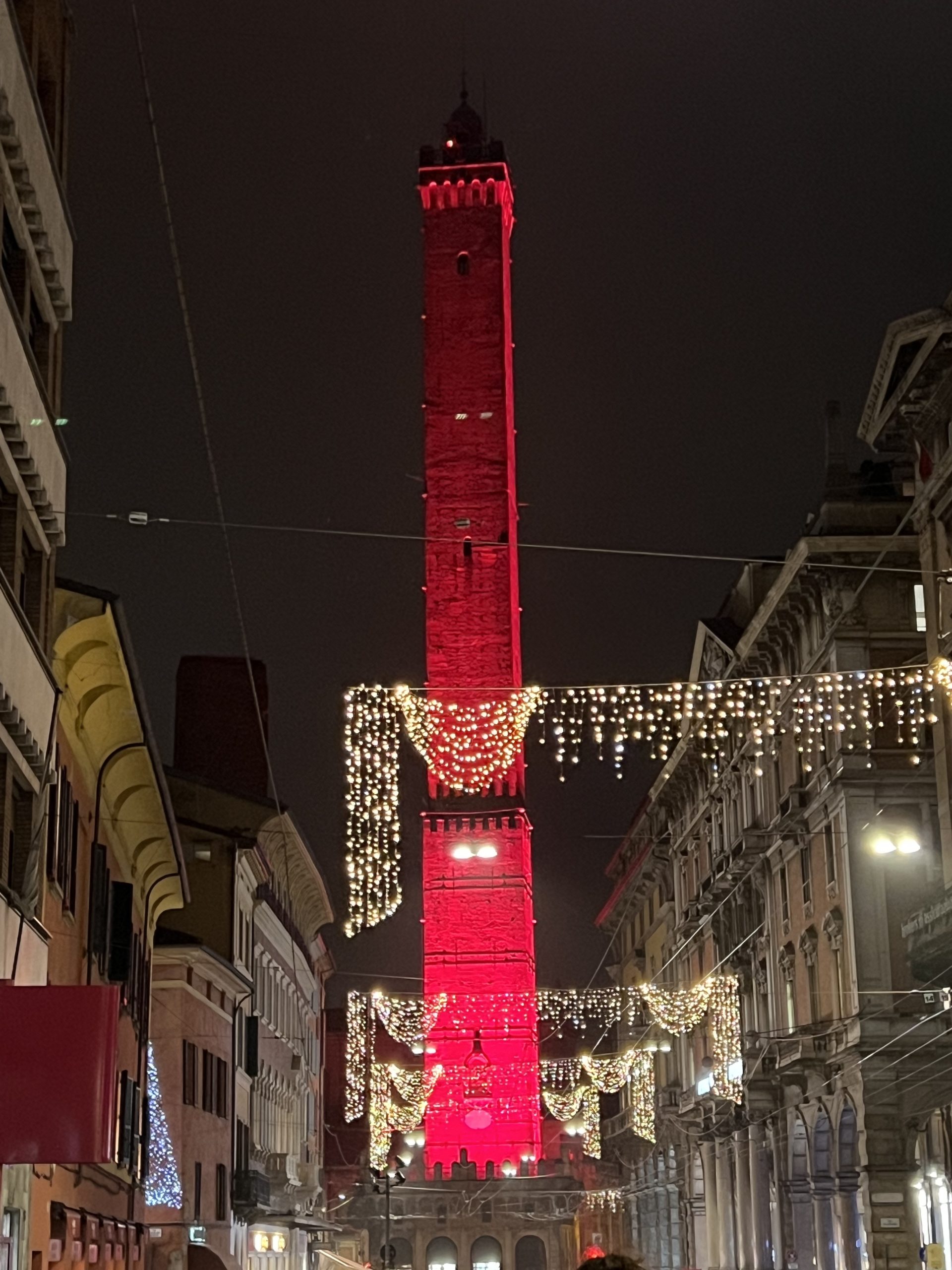 De noche, se recorta la silueta alargada y muy estilizada de la Torre Asinelli iluminada en rojo detrás de unas luces de navidad que cruzan la calle. Es Navidad 22